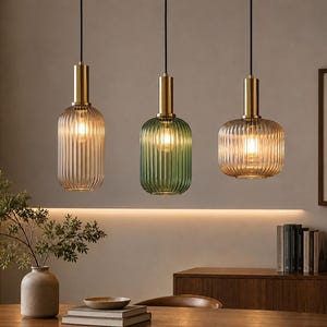 Könnte beinhalten: Drei Pendelleuchten mit gerippten Glasschirmen in klaren, grünen und bernsteinfarbenen Tönen. Jede Leuchte verfügt über eine zylindrische, messingfarbene Metallfassung an der Oberseite und hängt an einem dünnen schwarzen Kabel, das ein warmes Licht abgibt.
