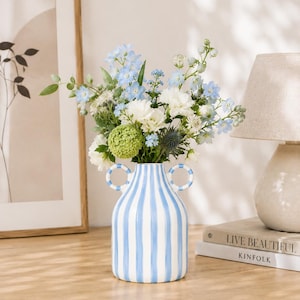 Könnte beinhalten: Eine Keramikvase mit rundem Körper und zwei kreisförmigen Henkeln, die mit vertikalen hellblauen und weißen Streifen versehen ist. Die Vase ist mit einem Strauß aus hellblauen Ritterspornen, weißen Nelken und grünen Hortensien gefüllt.
