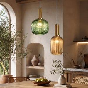 Può includere: Due lampade a sospensione con paralumi in vetro a coste, uno verde e uno ambrato, ciascuno dotato di un attacco cilindrico in metallo color ottone sulla parte superiore. Le luci sono sospese al soffitto tramite sottili cavi neri.