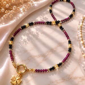 Op de afbeelding: Een kralenketting met afwisselend donkerrode en zwarte gefacetteerde kralen, afgewisseld met kleine goudkleurige metalen bolletjes en heldere gele kristallen accenten. De ketting heeft een goudkleurige knevelsluiting en een klaverblad-bedel.