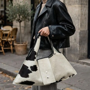 May include: A large hobo-style shoulder bag featuring a cow-print pattern with black and white patches. The bag is constructed from textured hair-on hide material, includes a central vertical panel with two silver-toned metal snap buttons, and black trim.