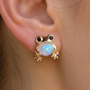 May include: A gold-toned stud earring shaped like a frog, featuring a round, iridescent white opal-like stone as the frog's body, with small black eyes and detailed webbed feet, securely fastened to the earlobe as a decorative piece of jewelry.