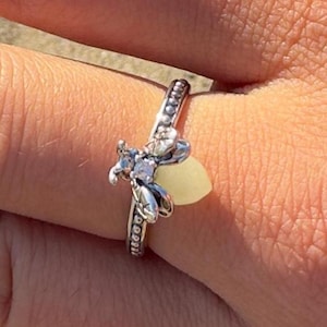 May include: A silver-toned ring featuring a bee-shaped centerpiece with a pale yellow, translucent, rounded stone body and small clear accent stones on the wings. The band has a textured, beaded edge design that encircles the finger.