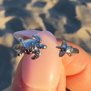 May include: A silver-toned open ring featuring a small sea turtle charm on one end and a tiny starfish charm on the other. The metal band is polished and reflective, showcasing detailed textures on both the turtle shell and the starfish points.