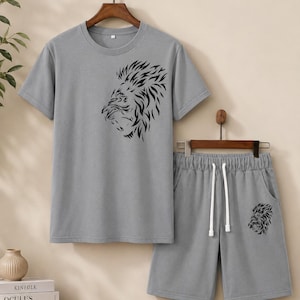 May include: A grey short-sleeved t-shirt and matching shorts set. The t-shirt features a black graphic of a lion's head on the chest. The shorts have an elastic waistband with white drawstrings and a smaller matching black lion head graphic.