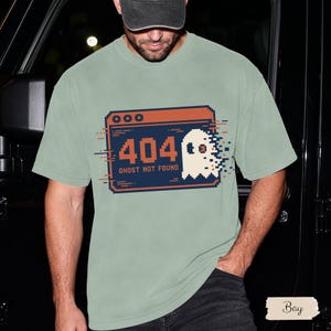 May include: A sage green t-shirt featuring a pixelated graphic of a browser window with the text 404 GHOST NOT FOUND. The design includes a white ghost icon with a glitch effect, rendered in orange and dark blue on the centre of the chest.