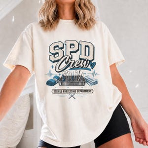 Könnte beinhalten: Ein cremefarbenes T-Shirt mit einem Aufdruck, der den Text SPD Crew und STERILE PROCESSING DEPARTMENT zeigt. Das Design umfasst eine Metallschale mit chirurgischen Instrumenten, ein blaues Bündel, Papierstapel, ein Herz und eine Schere.