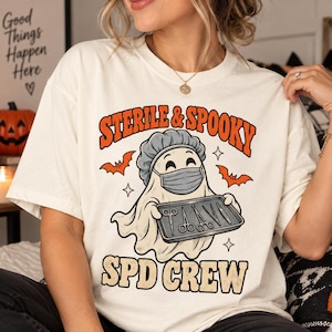 May include: An off-white t-shirt featuring a graphic of a ghost wearing a surgical cap and mask, holding a tray of medical instruments. The text reads "STERILE & SPOOKY" and "SPD CREW" in orange and black, accompanied by small orange bat illustrations.