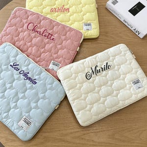 May include: Four rectangular quilted laptop sleeves in pastel yellow, pink, light blue, and off-white. Each sleeve features a cloud-patterned texture, a small white brand label, and custom embroidered names including arsilon, Charlotte, Los Angeles, and Murilo.
