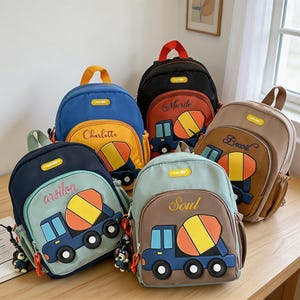 May include: Five backpacks in various colors including blue, black, navy, light green, and tan. Each bag features a front pocket with a graphic of a cement mixer truck and a name embroidered in cursive script, plus a small hanging charm.