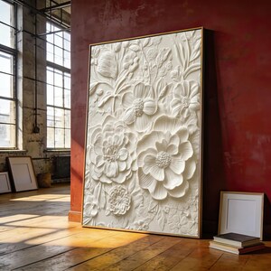 Op de afbeelding: Een rechthoekig wandkunstwerk met een witte, driedimensionale florale reliëfsculptuur in een dunne gouden lijst. Het kunstwerk toont een dichte compositie van diverse gebeeldhouwde bloemen, bloemblaadjes en bladeren met gedetailleerde texturen.