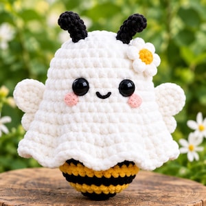 May include: A crocheted bee plushie wearing a white ghost costume. The bee has yellow and black stripes, while the ghost cover features black antennae, a white daisy flower, large black eyes, a small stitched smile, and pink cheeks.
