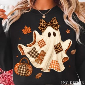 May include: A black long-sleeve sweatshirt featuring a central graphic of a white ghost adorned with a leopard print bow. The ghost is decorated with various patches including plaid, leopard print, and floral patterns, accompanied by a pumpkin and leaves.