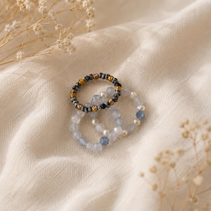 Könnte beinhalten: Ein Set aus drei Perlenringen. Ein Ring besteht aus gesprenkelten grauen und schwarzen Perlen mit goldfarbenen Metallzwischenstücken. Die anderen beiden Ringe bestehen aus durchscheinenden, hellblauen facettierten Perlen und kleinen weißen Kugeln.