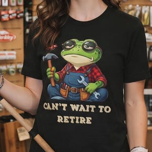 Può includere: Una t-shirt nera a maniche corte con la grafica di una rana verde che indossa occhiali da sole, una camicia a quadri rossi e una salopette blu, mentre tiene in mano un martello e una chiave inglese. Il testo "CAN'T WAIT TO RETIRE" è in giallo.
