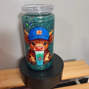 May include: A clear cylindrical tumbler filled with green glitter and liquid. The front features a graphic of a brown highland cow wearing a blue baseball cap with a logo, holding a small cup with a green drink and the text Seven Brew Coffee Company.