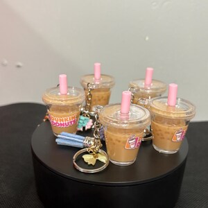 May include: A collection of miniature Dunkin Donuts iced coffee cup keychains. Each clear plastic cup is filled with a tan liquid, topped with a clear lid and a pink straw. The cups feature the Dunkin Donuts logo and include a metal ring.