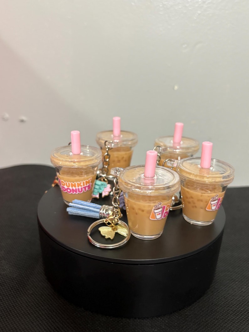 May include: A collection of miniature Dunkin Donuts iced coffee cup keychains. Each clear plastic cup is filled with a tan liquid, topped with a clear lid and a pink straw. The cups feature the Dunkin Donuts logo and include a metal ring.