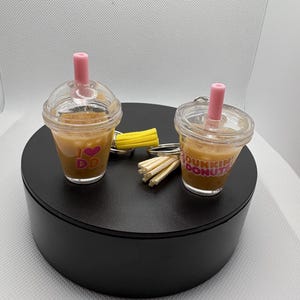 May include: Two miniature clear plastic cup keychains filled with a brown liquid, each featuring a pink straw and a lid. One cup displays the text I heart DD with a pink heart, while the other features the text Dunkin Donuts in pink and orange.