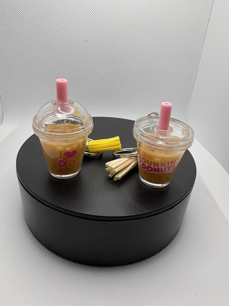 May include: Two miniature clear plastic cup keychains filled with a brown liquid, each featuring a pink straw and a lid. One cup displays the text I heart DD with a pink heart, while the other features the text Dunkin Donuts in pink and orange.