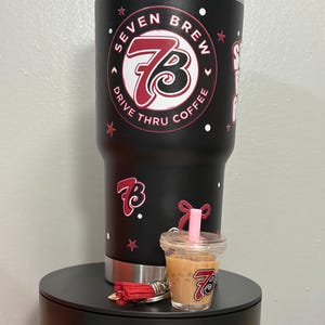May include: A matte black stainless steel tumbler with a clear lid and straw, featuring the Seven Brew Drive Thru Coffee logo in red, white, and black. A miniature iced coffee cup keychain with a pink straw and a red tassel is attached to the base.