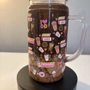 May include: A clear glass tumbler with a handle and lid, featuring a dark brown glittery upper section and a light brown liquid lower section. The exterior is decorated with a repeating pattern of Dunkin Donuts logos, coffee cups, donuts, and hearts.