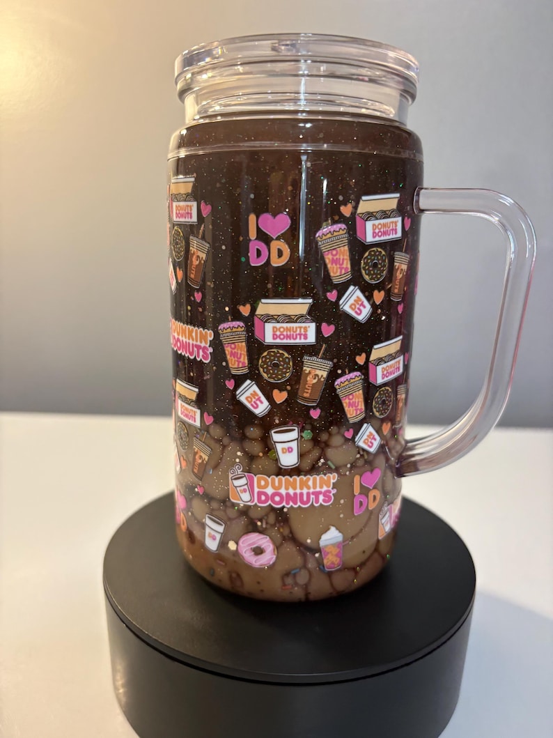 May include: A clear glass tumbler with a handle and lid, featuring a dark brown glittery upper section and a light brown liquid lower section. The exterior is decorated with a repeating pattern of Dunkin Donuts logos, coffee cups, donuts, and hearts.