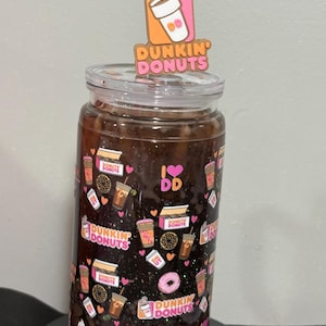 May include: A clear glass tumbler filled with a dark brown liquid, featuring a repeating pattern of Dunkin Donuts logos, coffee cups, donuts, and pink hearts. A clear straw is topped with a decorative Dunkin Donuts coffee cup acrylic topper.