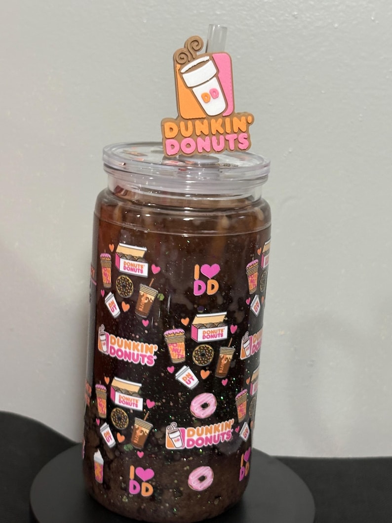 May include: A clear glass tumbler filled with a dark brown liquid, featuring a repeating pattern of Dunkin Donuts logos, coffee cups, donuts, and pink hearts. A clear straw is topped with a decorative Dunkin Donuts coffee cup acrylic topper.