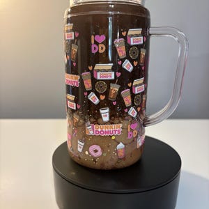 May include: A clear glass tumbler with a handle and lid, featuring a dark brown liquid base and a pattern of Dunkin Donuts logos, coffee cups, donuts, and pink hearts. The design includes the text I LOVE DD and DUNKIN DONUTS throughout.