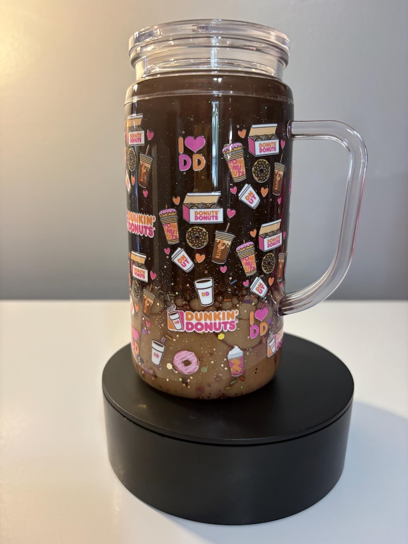 May include: A clear glass tumbler with a handle and lid, featuring a dark brown liquid base and a pattern of Dunkin Donuts logos, coffee cups, donuts, and pink hearts. The design includes the text I LOVE DD and DUNKIN DONUTS throughout.