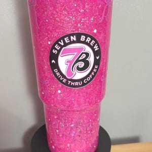 May include: A bright pink glitter tumbler with a clear lid, featuring the Seven Brew Drive Thru Coffee logo in the center. The exterior surface is decorated with small pink bow icons scattered throughout the vibrant, sparkling pink liquid.