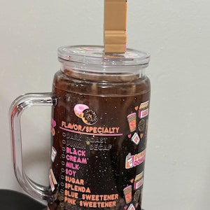 May include: A clear glass tumbler with a handle and a lid featuring a brown donut-shaped straw topper. The exterior is decorated with a Dunkin' Donuts menu list including options like dark roast, black, cream, milk, soy, sugar, and various sweeteners.
