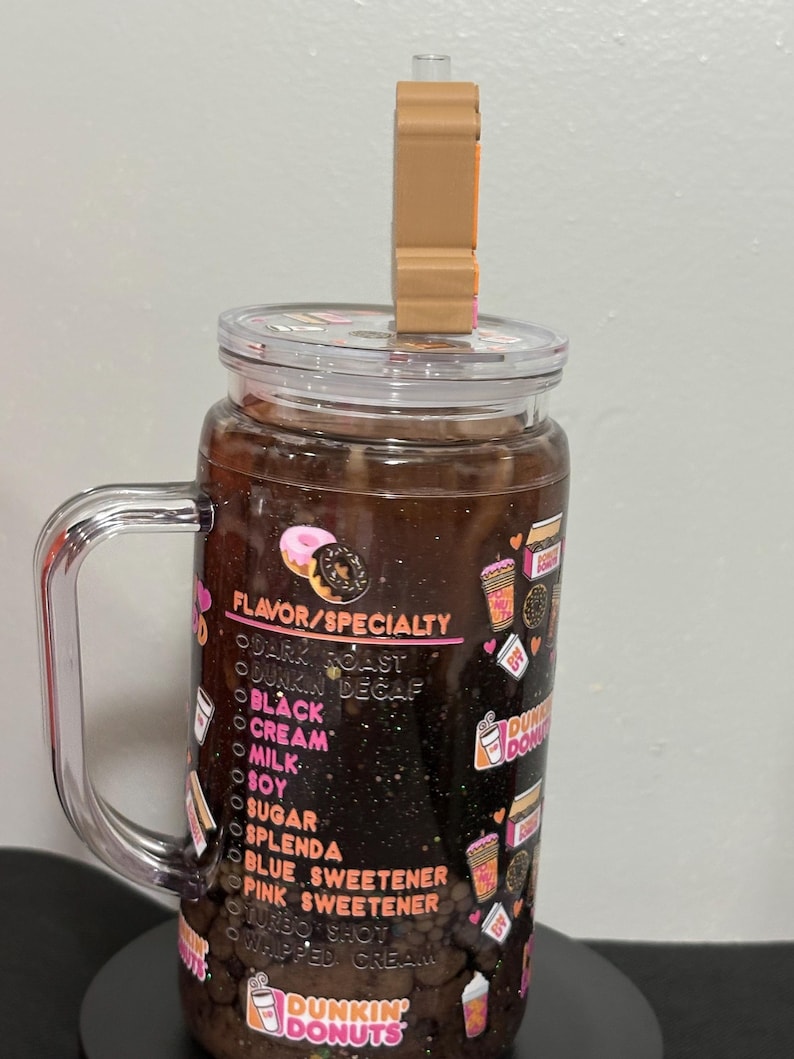 May include: A clear glass tumbler with a handle and a lid featuring a brown donut-shaped straw topper. The exterior is decorated with a Dunkin' Donuts menu list including options like dark roast, black, cream, milk, soy, sugar, and various sweeteners.