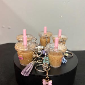 May include: A collection of miniature iced coffee cup keychains, each featuring a clear plastic cup filled with a tan liquid, a pink straw, and a metal keyring. Some keychains include a small lavender tassel or a pink bow charm attached.