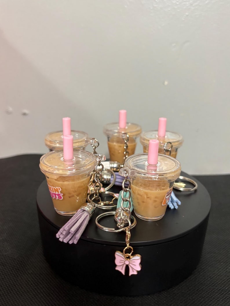 May include: A collection of miniature iced coffee cup keychains, each featuring a clear plastic cup filled with a tan liquid, a pink straw, and a metal keyring. Some keychains include a small lavender tassel or a pink bow charm attached.