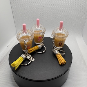 May include: Three miniature iced coffee cup keychains with clear plastic lids and pink straws. Each cup contains a tan liquid and features a small graphic print. Attached to each silver-toned metal keyring is a bright yellow decorative tassel.