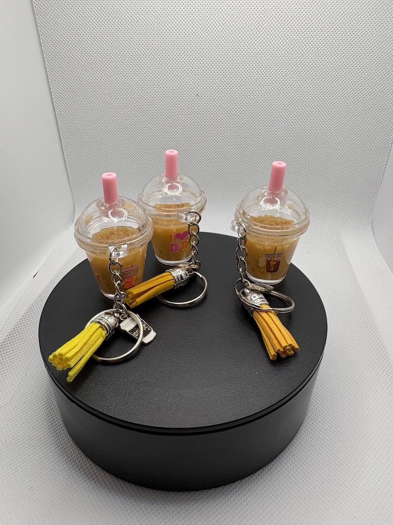 May include: Three miniature iced coffee cup keychains with clear plastic lids and pink straws. Each cup contains a tan liquid and features a small graphic print. Attached to each silver-toned metal keyring is a bright yellow decorative tassel.