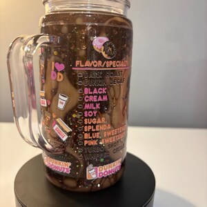 May include: A clear acrylic tumbler with a handle and lid, filled with a dark brown liquid and glitter. The exterior features Dunkin Donuts branding, a list of flavor and specialty options, and small donut graphics printed in pink, white, and orange.