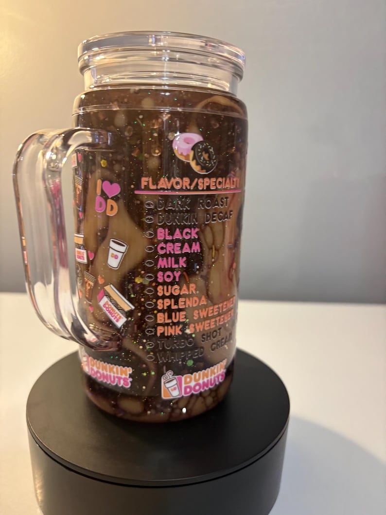 May include: A clear acrylic tumbler with a handle and lid, filled with a dark brown liquid and glitter. The exterior features Dunkin Donuts branding, a list of flavor and specialty options, and small donut graphics printed in pink, white, and orange.