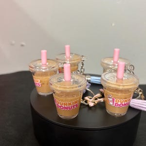 May include: A collection of miniature clear plastic cup keychains filled with a tan liquid resembling iced coffee. Each cup features a pink straw, a clear lid, and the Dunkin Donuts logo printed in orange and pink on the front surface.