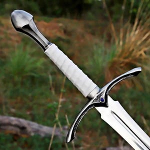 Op de afbeelding: Een zwaard met een gepolijste zilverkleurige metalen pommel, een wit omwikkelde greep en een stootplaat met een kleine blauwe ronde inleg in het midden. Het lemmet is gemaakt van reflecterend zilverkleurig metaal met een centrale richel.