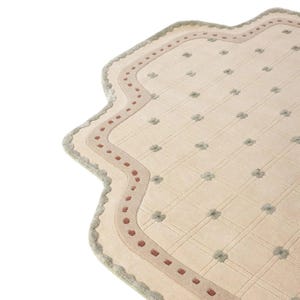 May include: A cream-colored area rug with a scalloped edge and a grid pattern. The rug features small, grey, four-petaled floral motifs at each grid intersection, a border with a row of small, rust-colored squares, and a light grey trim.