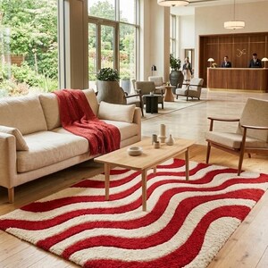 May include: A rectangular area rug featuring a repeating wavy pattern in deep red and cream white. The rug has a thick, textured pile and is positioned on a light-colored wooden floor in front of a light beige sofa with a red throw blanket.