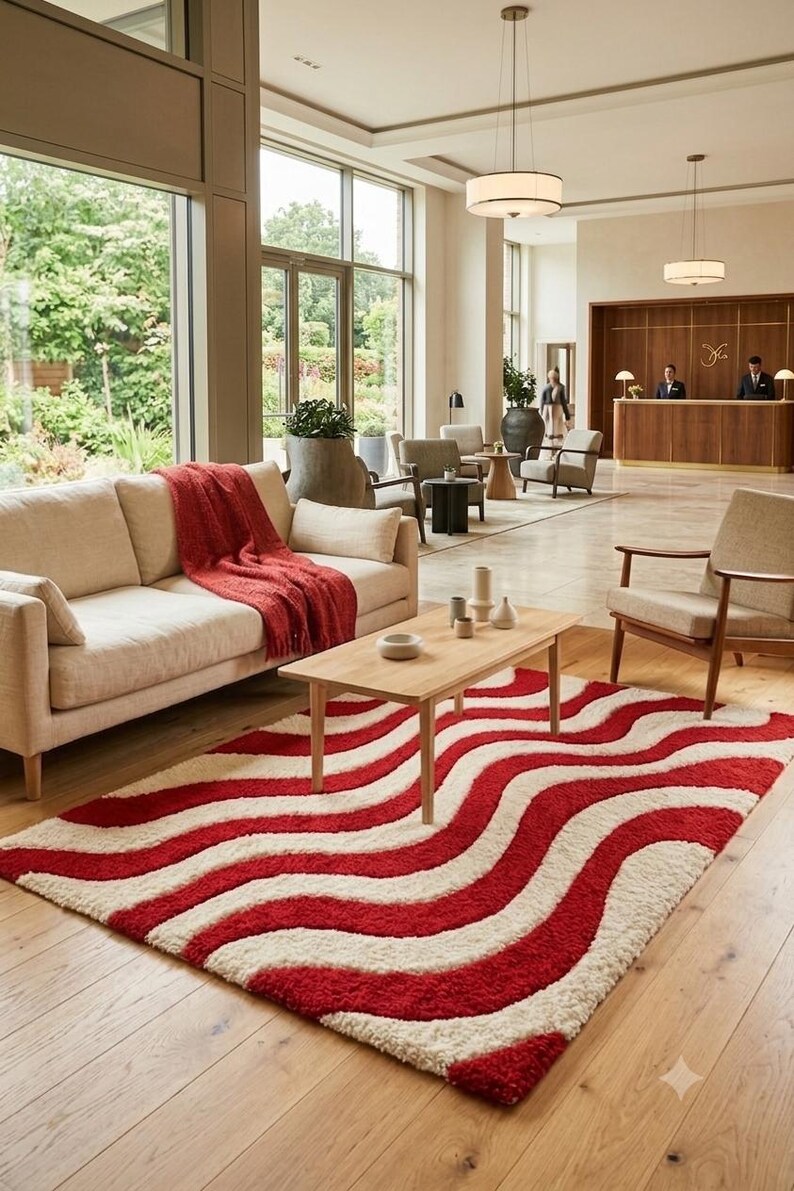 May include: A rectangular area rug featuring a repeating wavy pattern in deep red and cream white. The rug has a thick, textured pile and is positioned on a light-colored wooden floor in front of a light beige sofa with a red throw blanket.