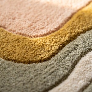May include: A tufted rug featuring a series of curved, wavy bands in soft pastel colors. The texture is thick and plush, displaying distinct layers of light pink, mustard yellow, sage green, and cream white fibers arranged in a flowing pattern.