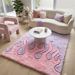 May include: A rectangular area rug featuring a plush, shaggy texture with a repeating flame pattern in alternating shades of soft pastel pink and light lavender. The rug is positioned on a light wood floor beneath a white marble circular coffee table.