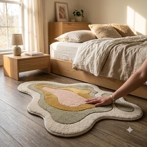 May include: An irregular, organic-shaped area rug featuring layered, wavy bands of color including cream, sage green, mustard yellow, and soft pink. The rug has a thick, tufted, textured pile surface and is positioned on a dark wood floor.