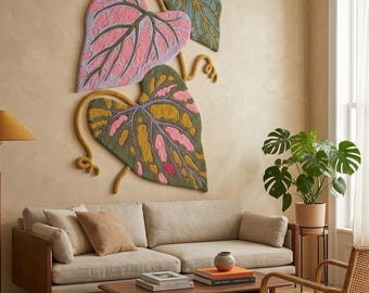 Puede incluir: Un gran tapiz de pared texturizado que presenta tres hojas estilizadas de gran tamaño en tonos rosa, lavanda, verde oliva y amarillo mostaza. El arte textil con mechones muestra patrones de venas intrincados y tallos curvos en la pared.