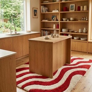 May include: A rectangular area rug featuring a bold, wavy pattern of thick, textured red and cream-colored stripes. The rug is positioned on a light-colored wooden floor, extending underneath a wooden kitchen island in a bright, open-plan space.